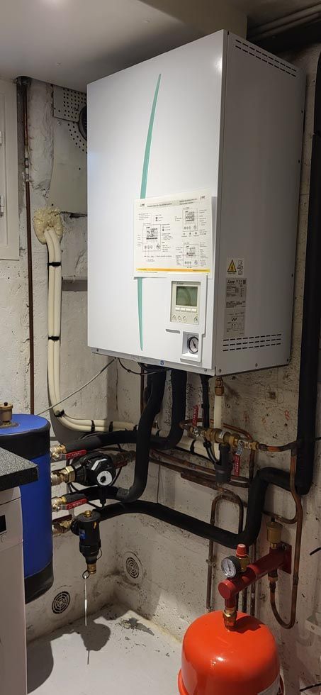 Une chaudière blanche avec des touches de vert, fixée au mur d'un sous-sol, reliée à des tuyaux et des réservoirs.