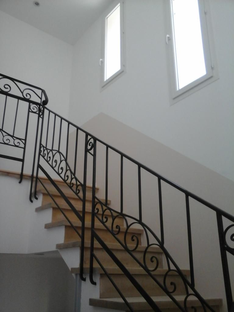 Escalier avec une rampe en fer forgé, des marches claires et deux hautes fenêtres sur un mur blanc.