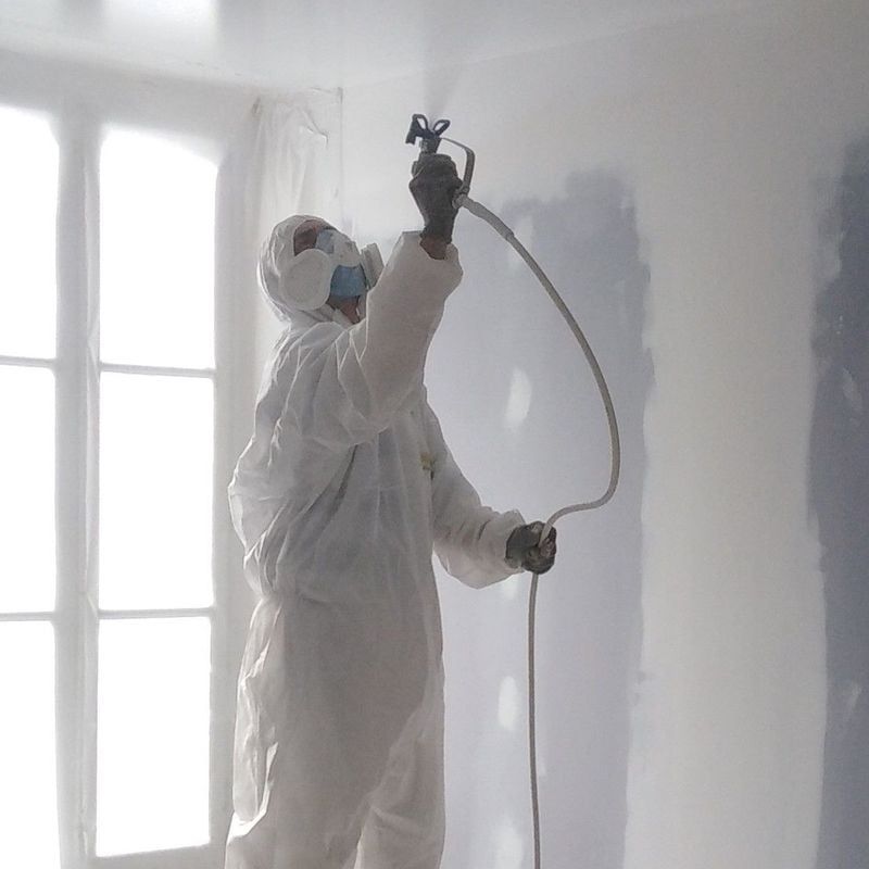 Une personne en combinaison de protection vaporise de la peinture sur un mur à l'intérieur.