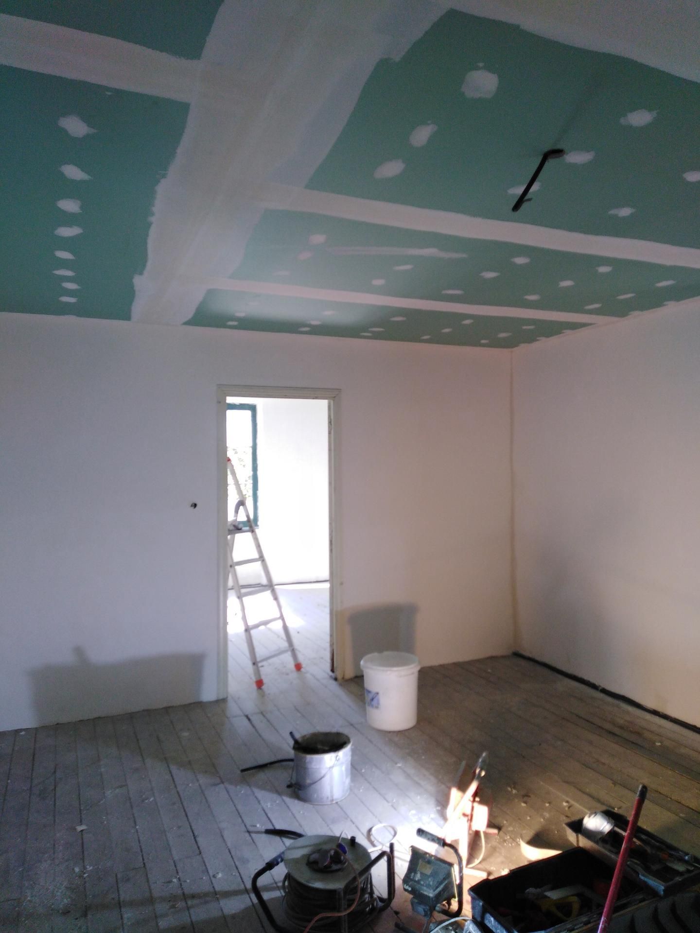 Pièce en cours de rénovation ; plafond en plaques de plâtre, murs blancs, porte ouverte, outils sur le plancher en bois.