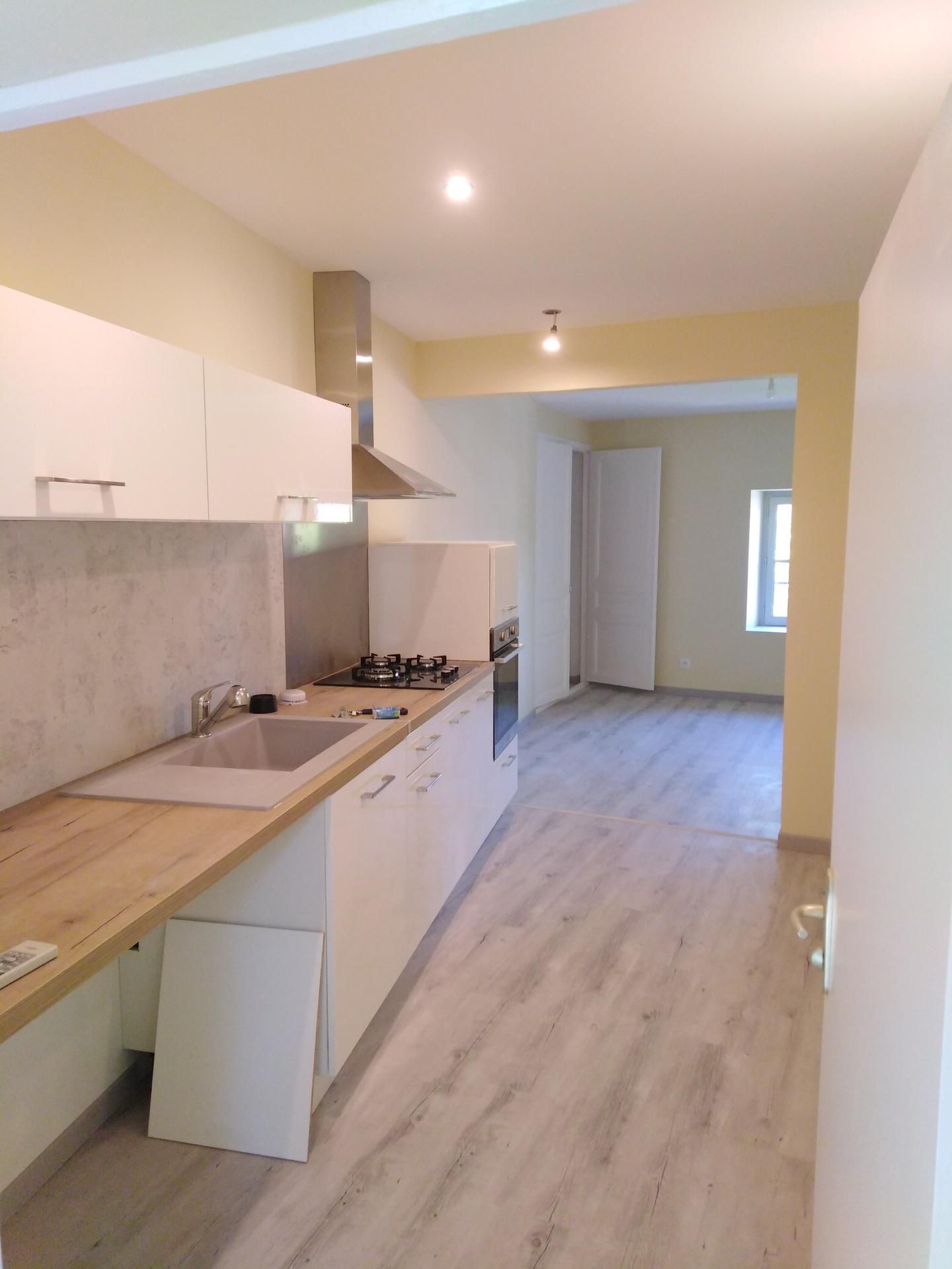 Cuisine moderne avec placards blancs, plan de travail en bois et crédence gris clair, donnant sur une autre pièce.