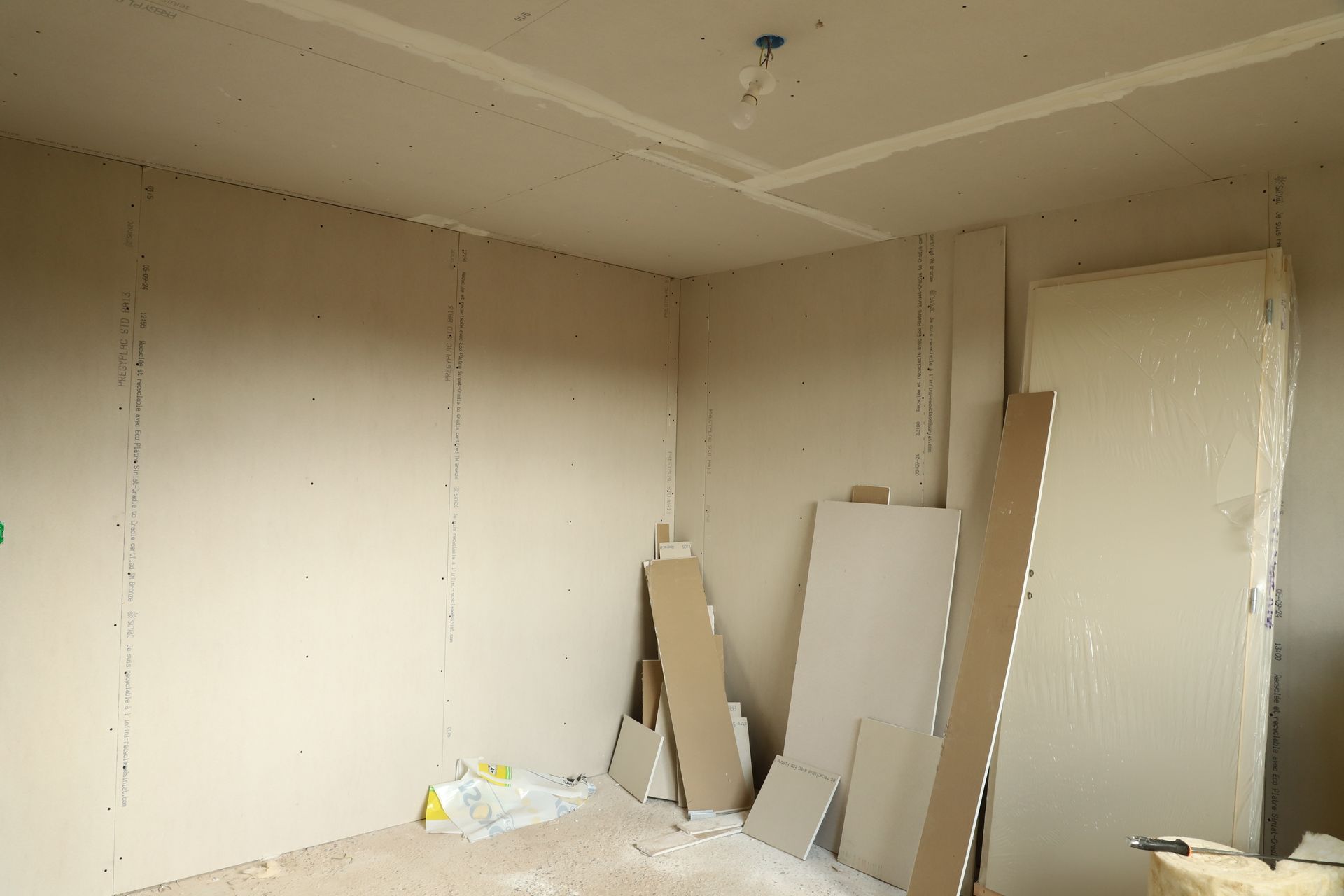 Intérieur d'une pièce en construction, avec des plaques de plâtre installées sur les murs et le plafond ; quelques plaques de plâtre sont empilées dans un coin.