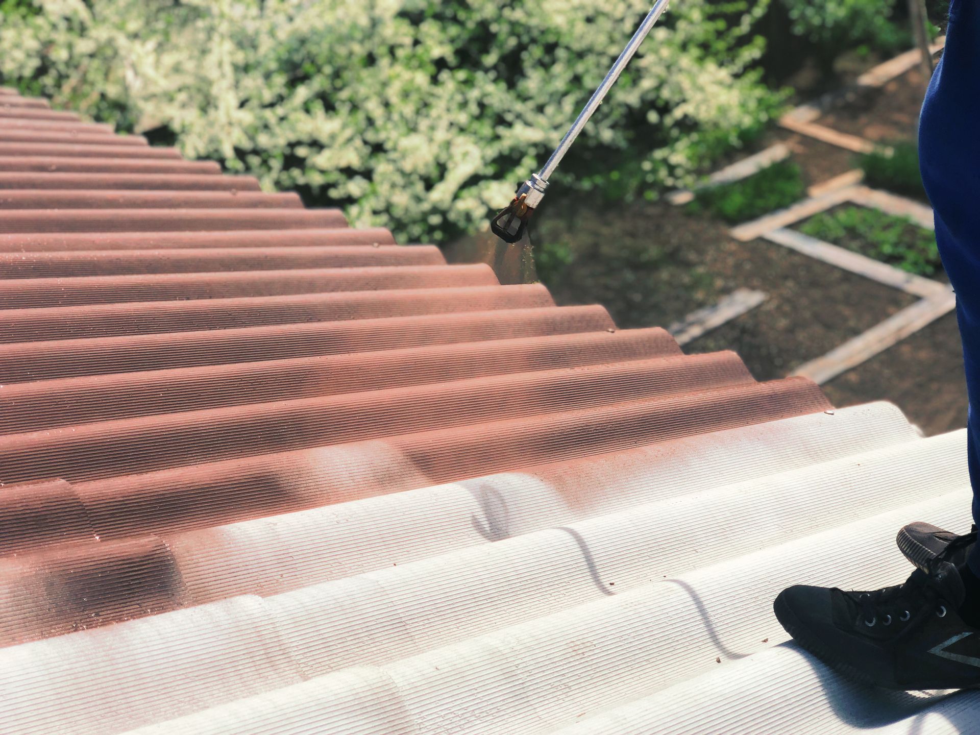 Une personne nettoie avec un jet à haute pression un toit de tuiles rouges, de haut en bas.