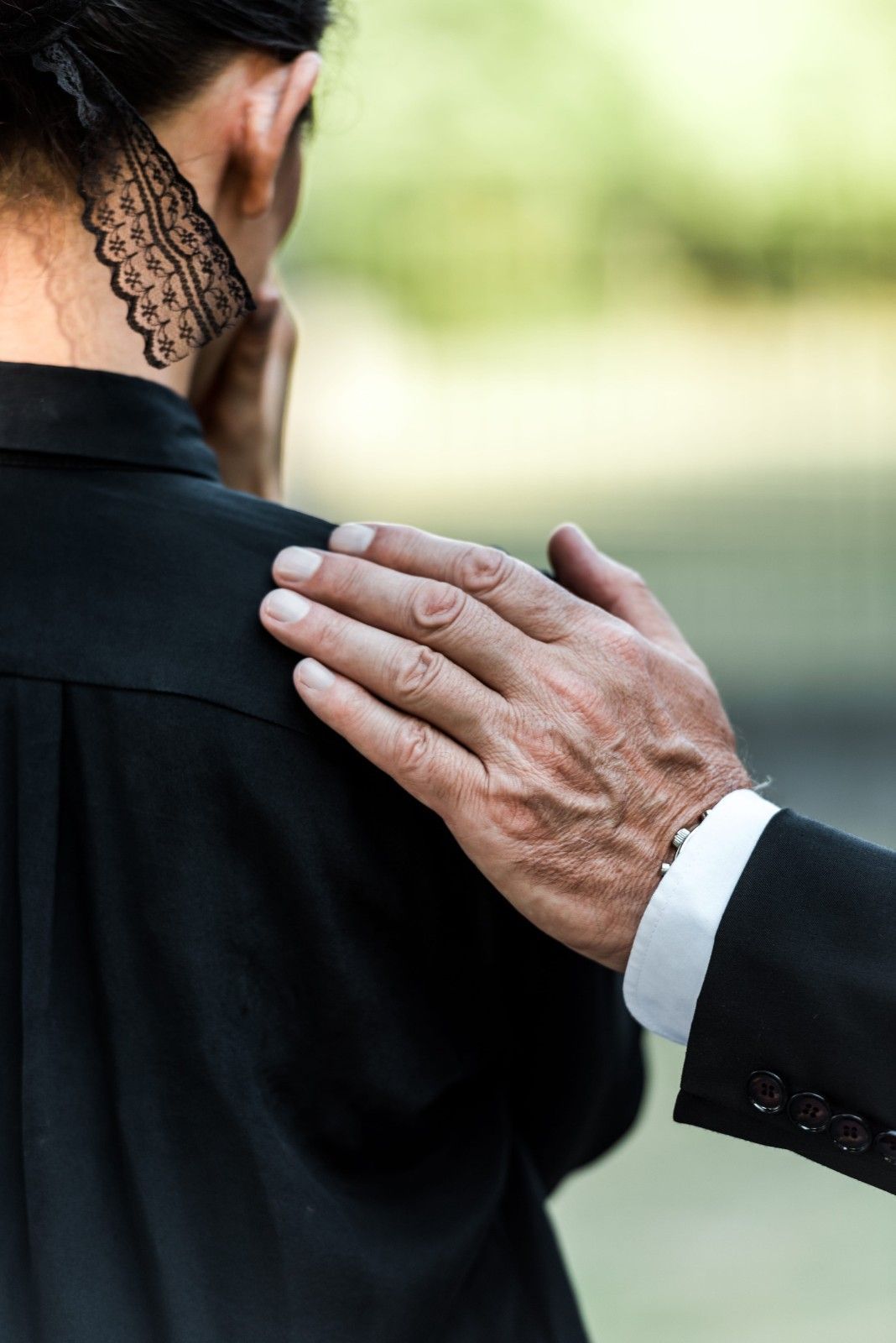 Una mano, asomada a la manga de un traje negro, descansa sobre el hombro de una persona que lleva una camisa negra.