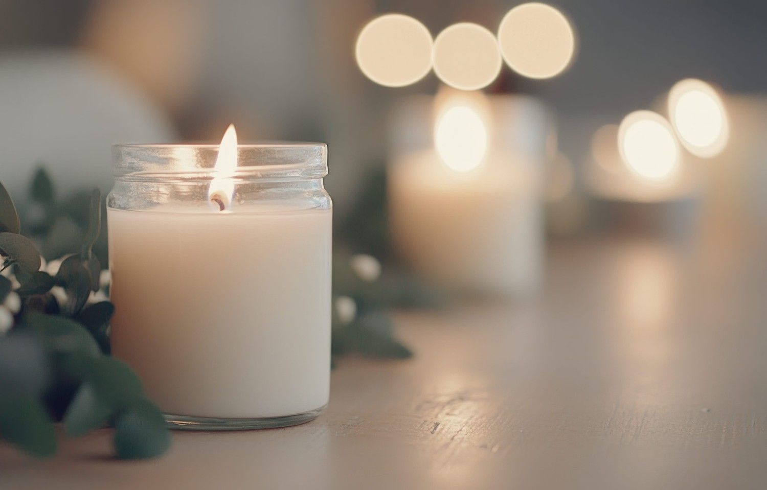 Una vela encendida en un frasco de vidrio reposa sobre una superficie junto a follaje verde, con velas ligeramente borrosas.