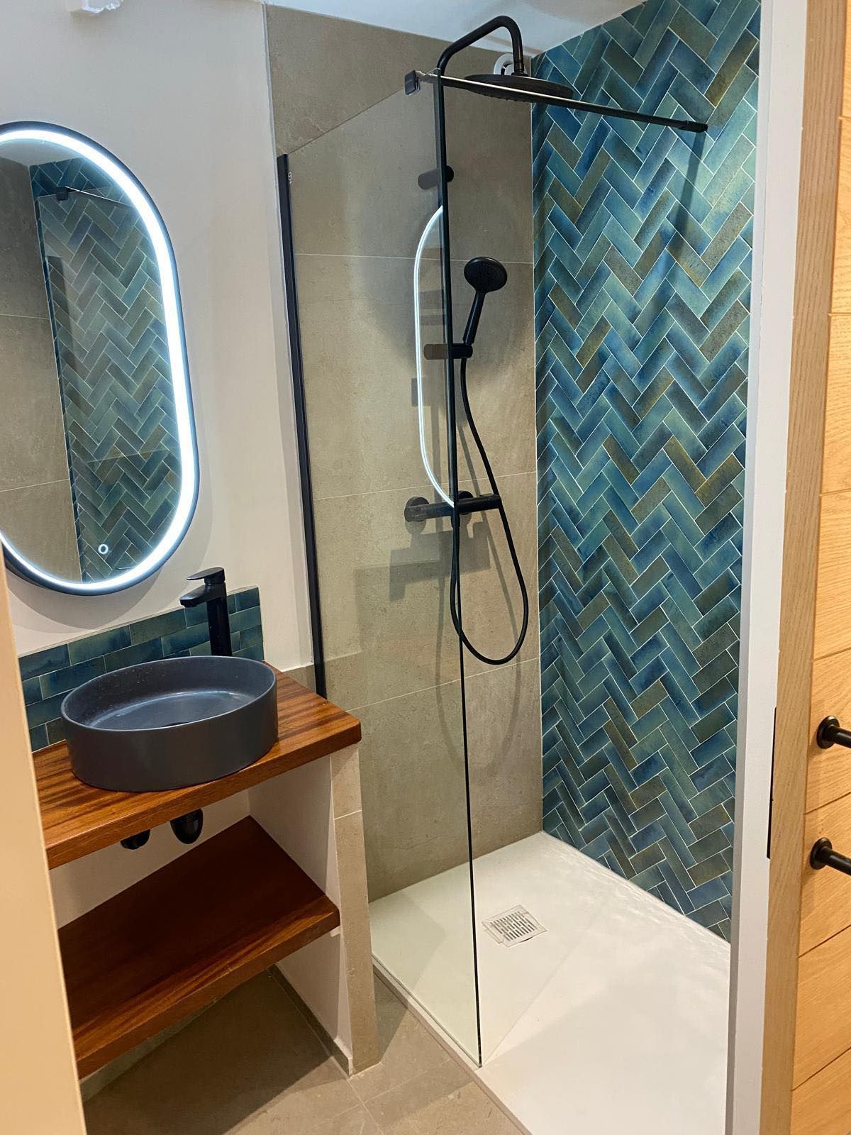 Salle de bains avec douche à l'italienne, carrelage turquoise, robinetterie noire et meuble lavabo en bois avec lavabo gris.