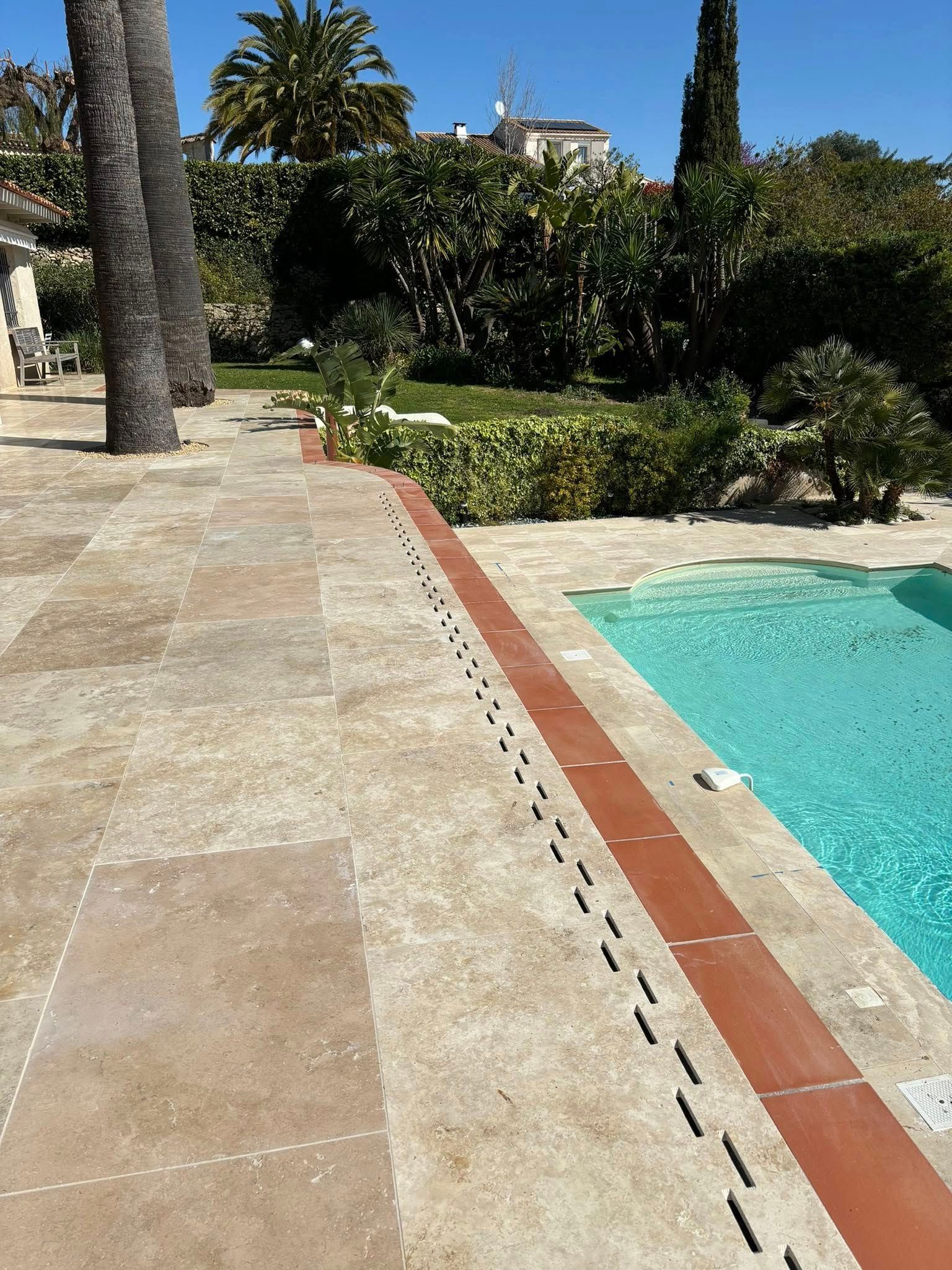 Vue sur la piscine avec carrelage en pierre, bordure en briques et eau bleue. Palmiers et verdure en arrière-plan.