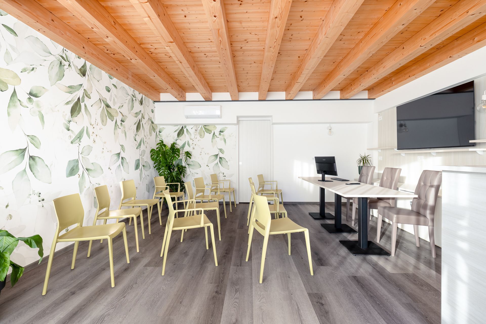 Una stanza moderna con travi in ​​legno, carta da parati floreale, sedie dai colori chiari  e una scrivania con monitor.
