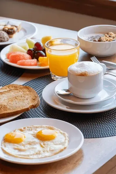 Una colazione imbandita con uova all'occhio di bue, pane tostato, frutta, succo d'arancia, un cappuccino e cereali.