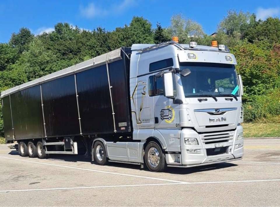 Un semi-remorque MAN argenté avec une remorque noire est stationné sur le trottoir près des arbres.