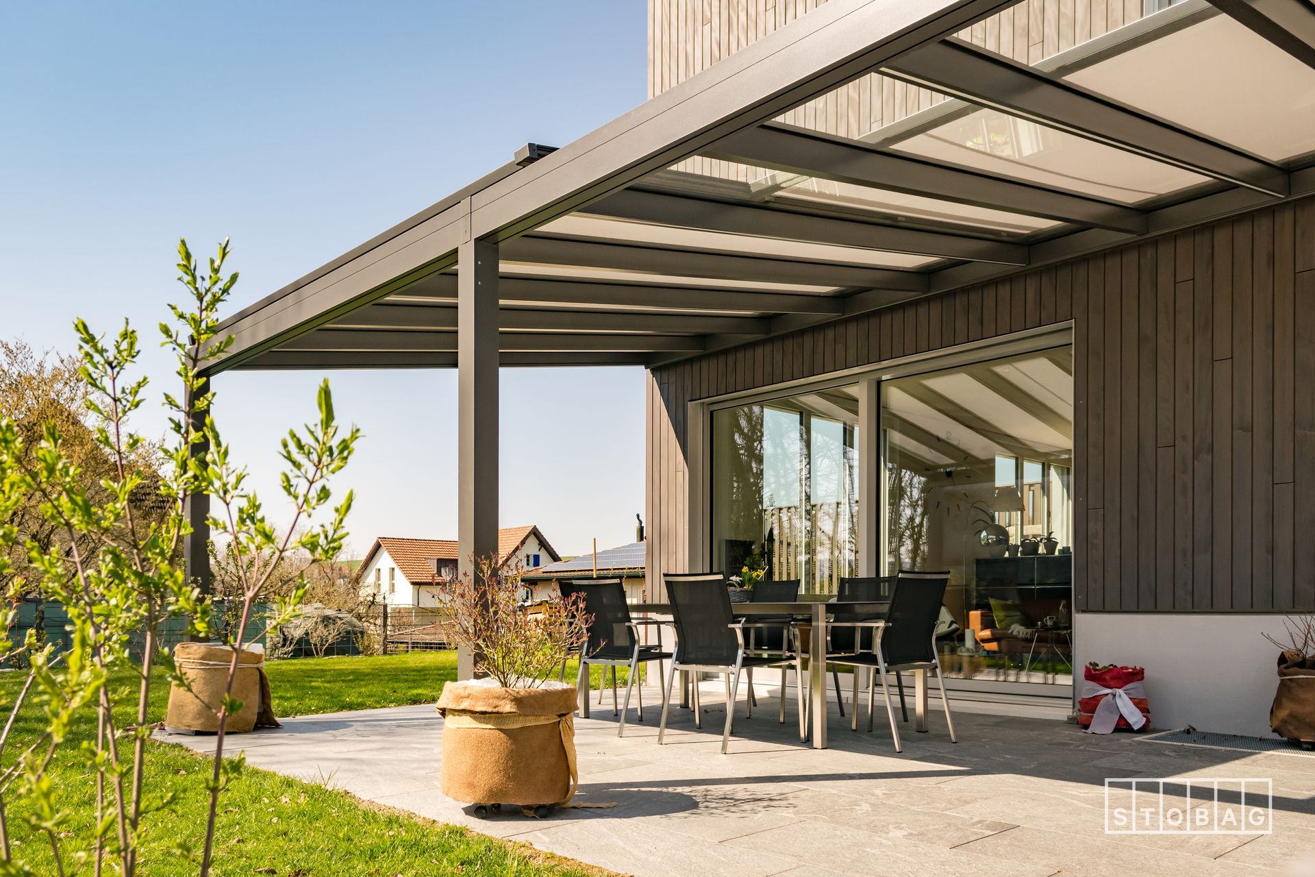 Patio esterno con tavolo da pranzo e sedie, sotto un pergolato coperto attaccato a un edificio.