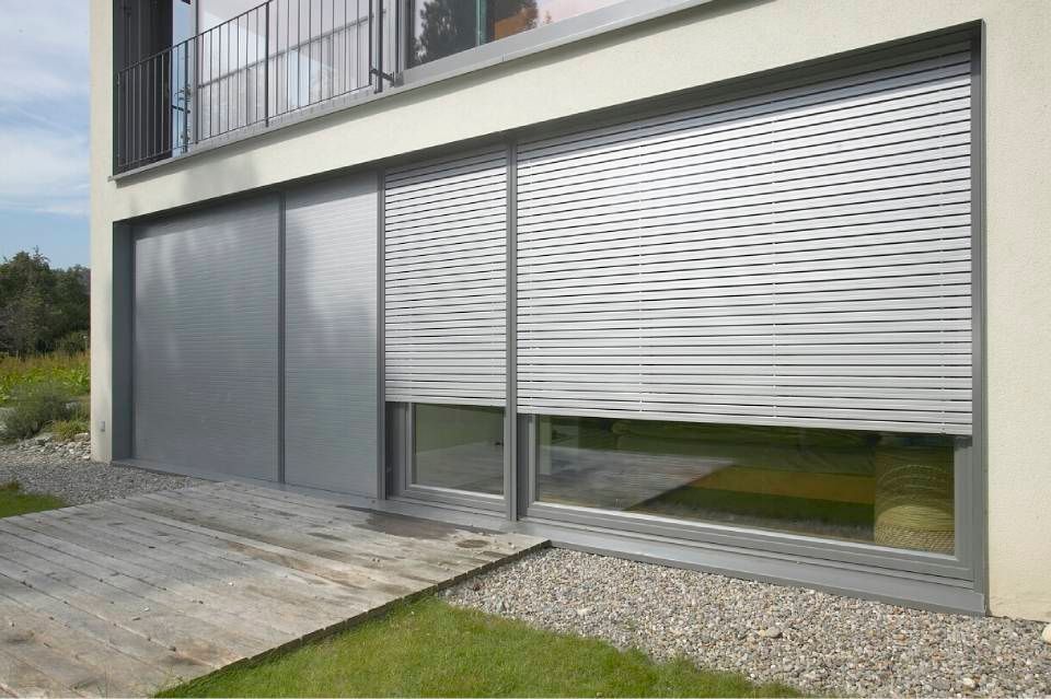 Edificio moderno con persiane grigie chiuse sopra una finestra e una porta scorrevole, accanto a una terrazza in legno.
