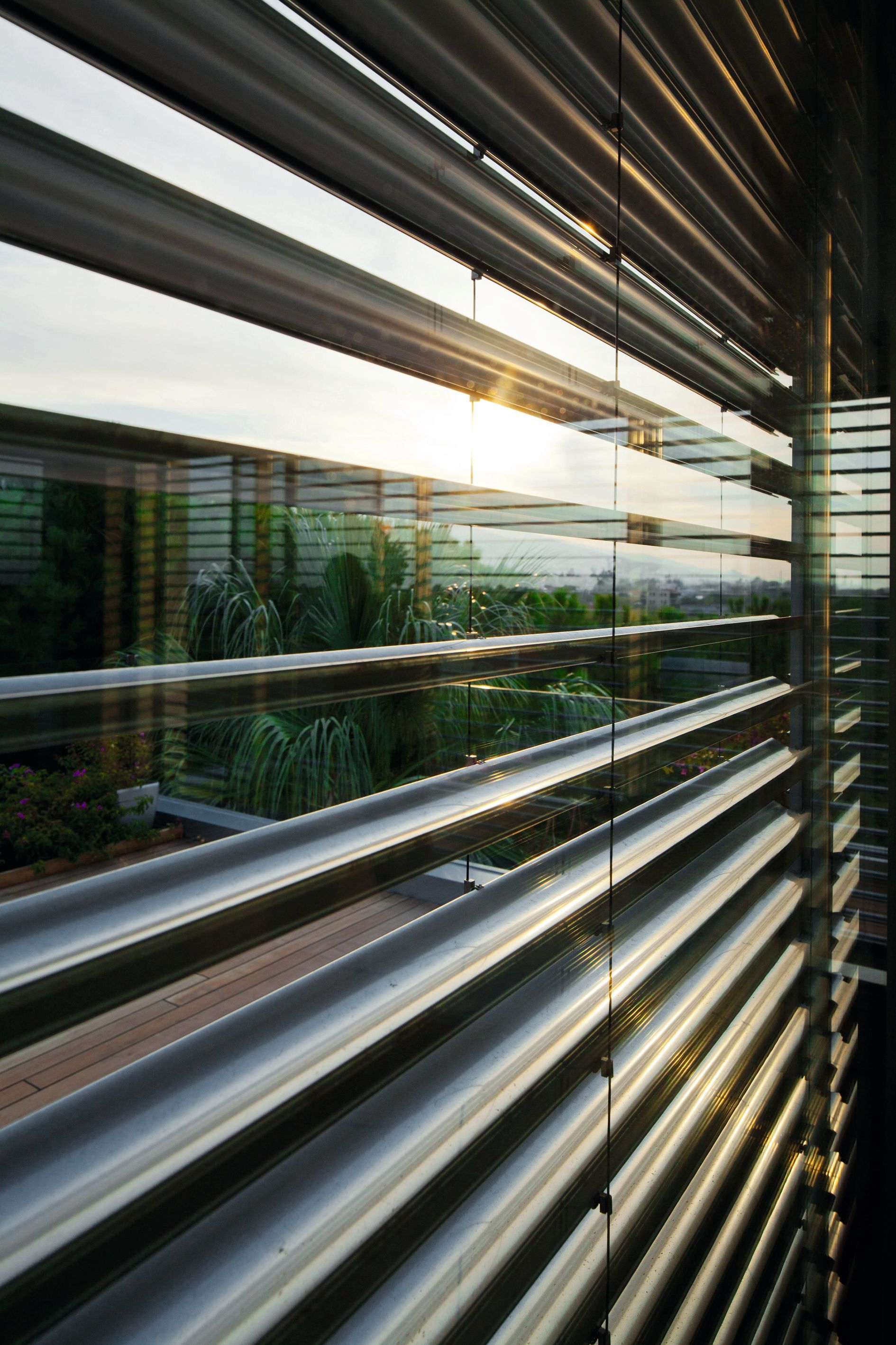 Persiane metalliche che riflettono la luce del sole, con vista sul verde giardino pensile e sul paesaggio urbano.