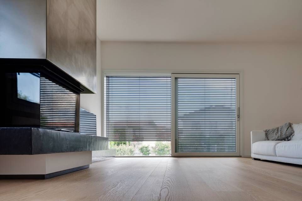 Soggiorno con porta scorrevole in vetro, tende orizzontali, pavimento in legno chiaro, divano bianco e camino.