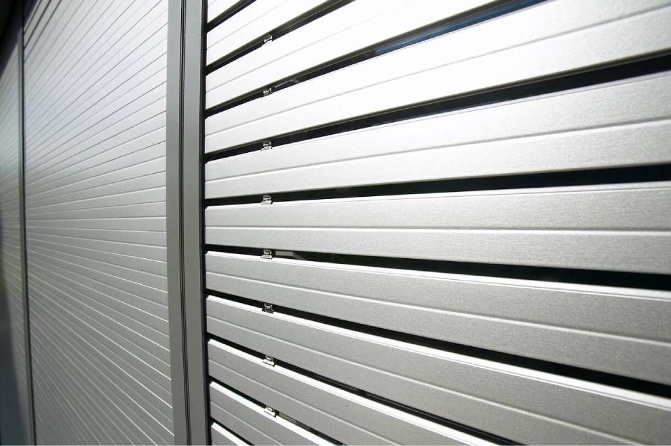 Primo piano di una persiana in metallo argentato; parzialmente aperta, rivela le stecche orizzontali e il muro.