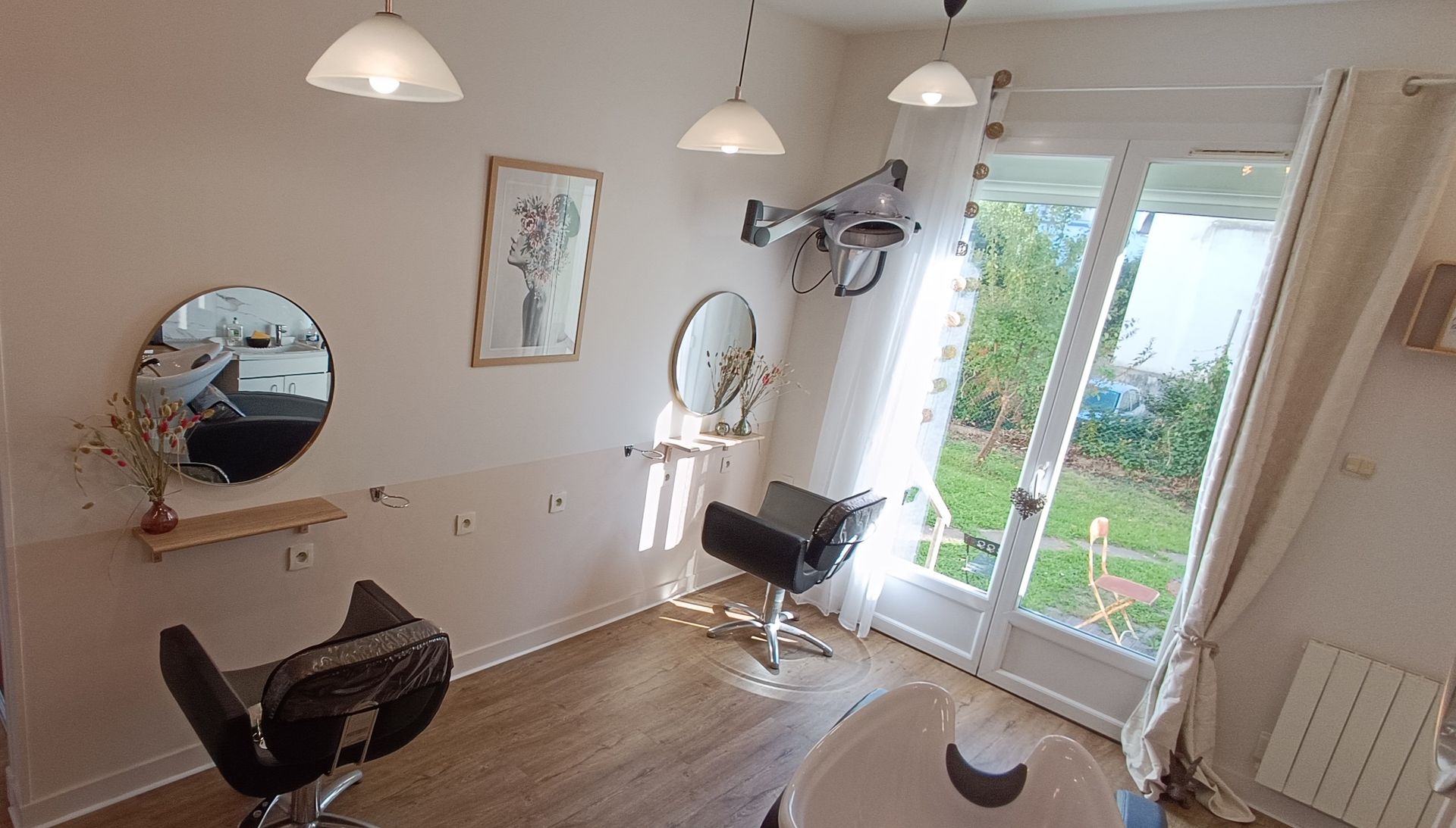 L'intérieur d'un salon de coiffure avec deux fauteuils de coiffure, des miroirs circulaires, des suspensions lumineuses et une vue sur une cour gazonnée à travers les portes.