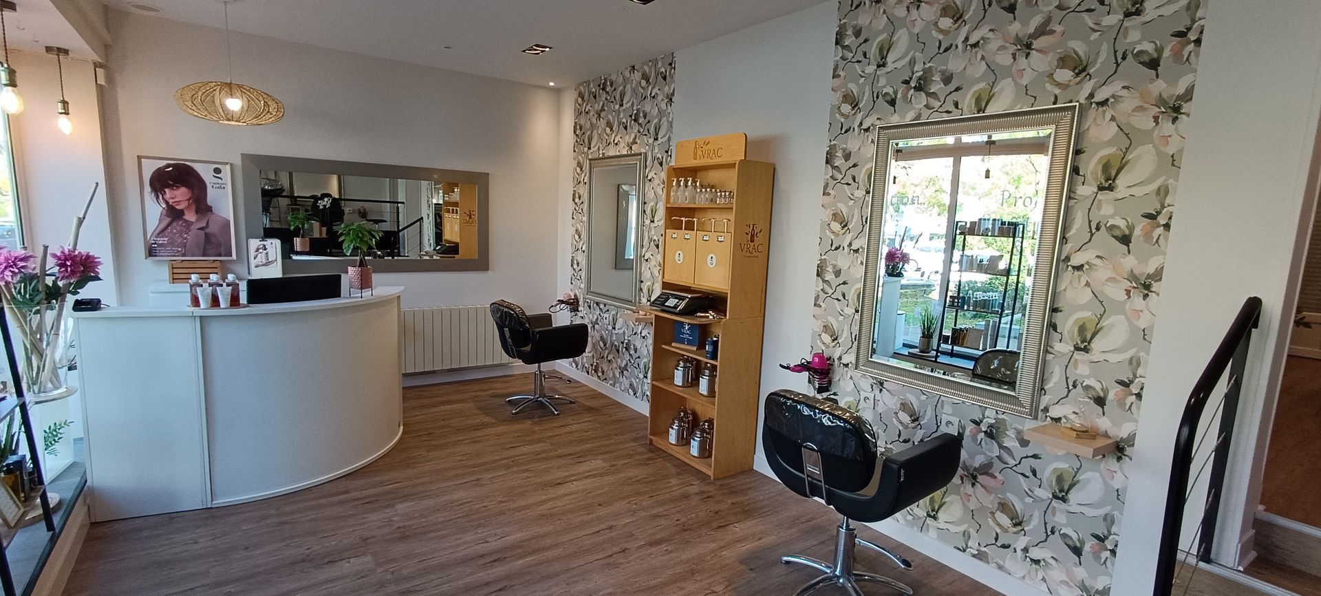Intérieur d'un salon de coiffure avec un comptoir d'accueil blanc incurvé, deux fauteuils de coiffure, une étagère de présentation de produits et un papier peint à motifs floraux.