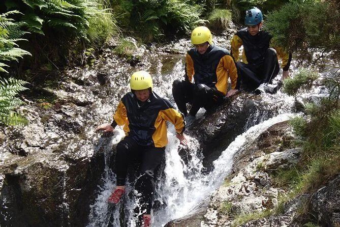 Three people in wetsuits and helmets navigate a rocky stream, water splashing.