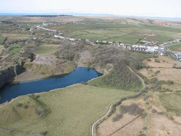 Aerial view of a quarry lake surrounded by green fields and a small town in the distance under a blue sky.