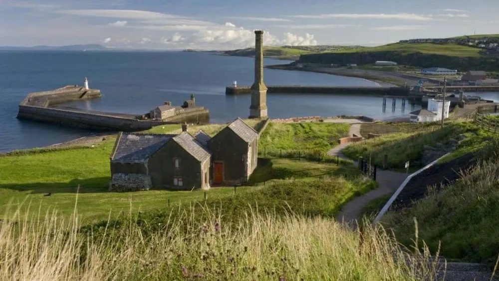 Coastal view of a harbor with stone buildings, a tall chimney, a grassy hill, and the sea.