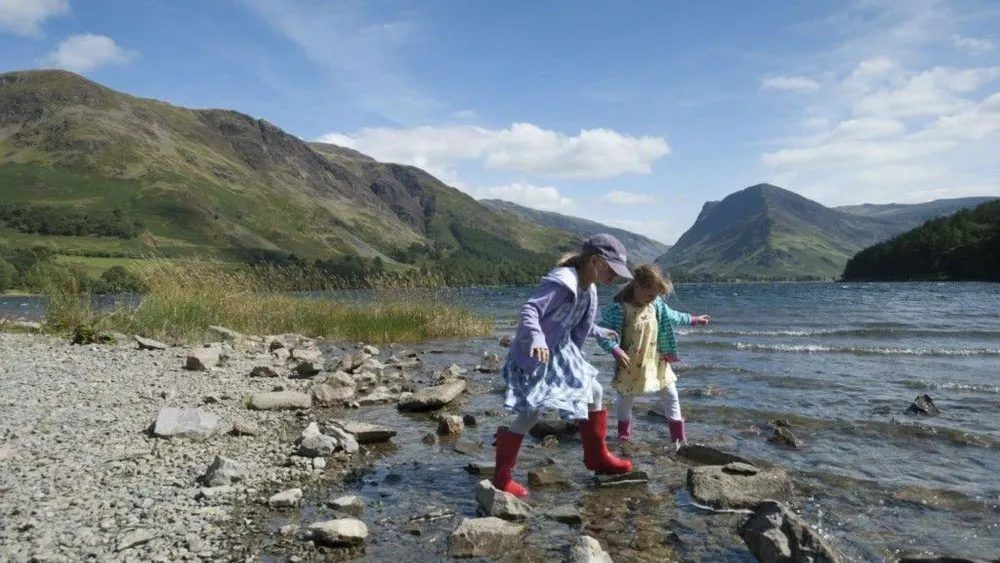 Two children in rain boots play at water's edge of a lake with mountains in the background.