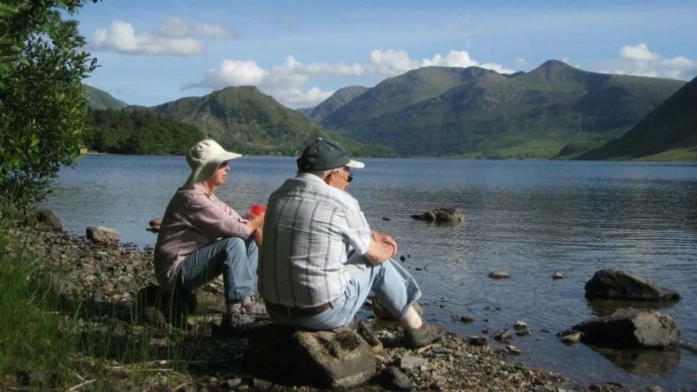 Two people sit by a lake, looking at mountains in the distance under a blue sky.
