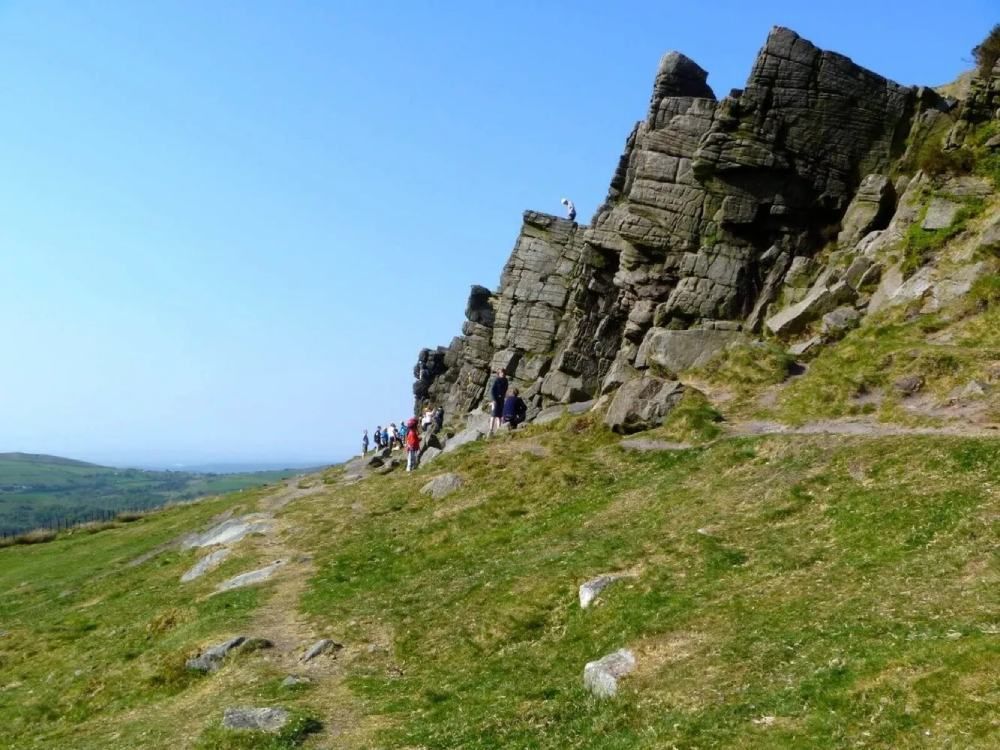 A rocky cliff face next to a grassy hill with hikers on a clear, sunny day.