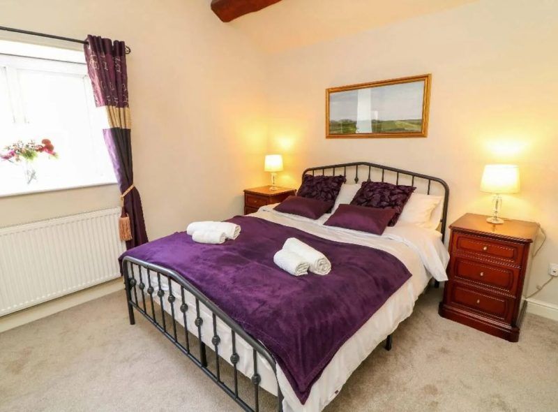 Bedroom with purple bedding, metal bed frame, two bedside tables with lamps, and a window with curtains.