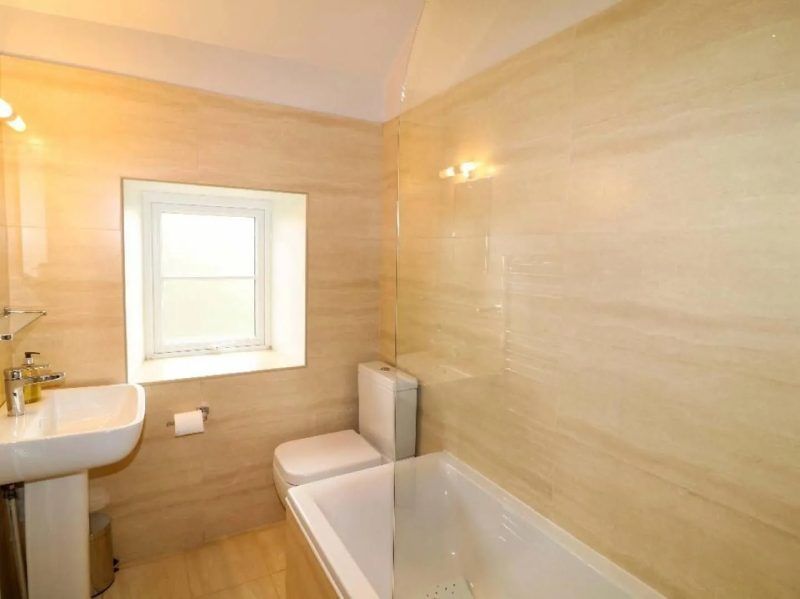 Bathroom with a pedestal sink, toilet, tub, and window. Beige tile walls, white fixtures, and natural light.