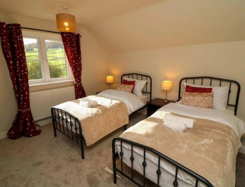 Two twin beds with beige blankets in a light-filled bedroom, red curtains at the window.