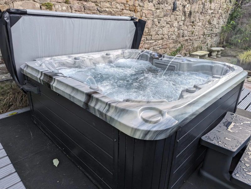 Hot tub with bubbling water and partially open cover, set on a deck next to a stone wall.
