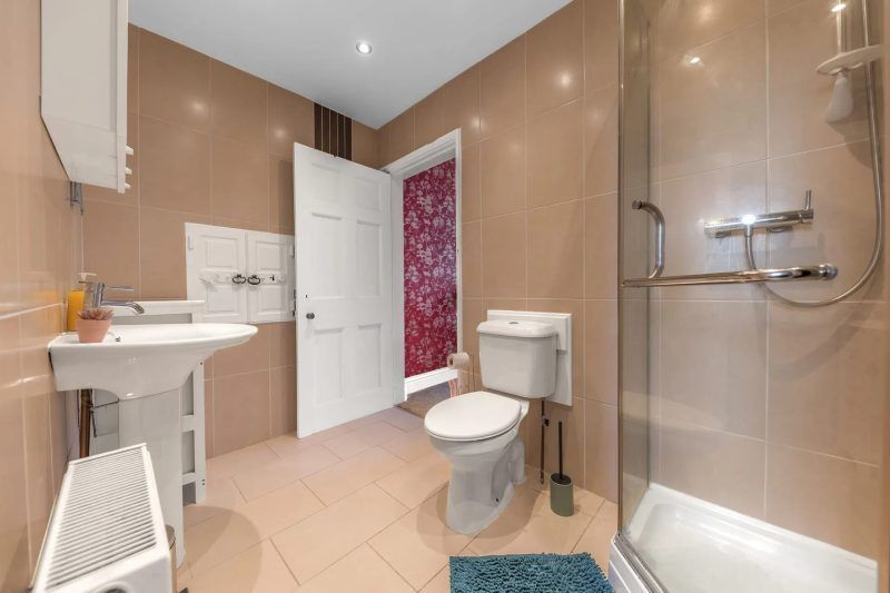Bathroom with beige tile walls and floor. Includes a toilet, sink, shower, and open doorway leading to a red patterned wall.