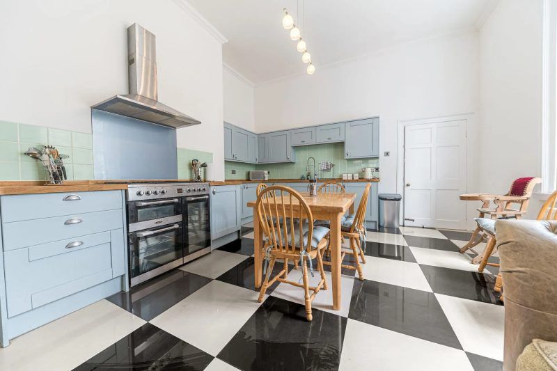 Kitchen with blue cabinets, wooden table and chairs, black and white checkered floor.