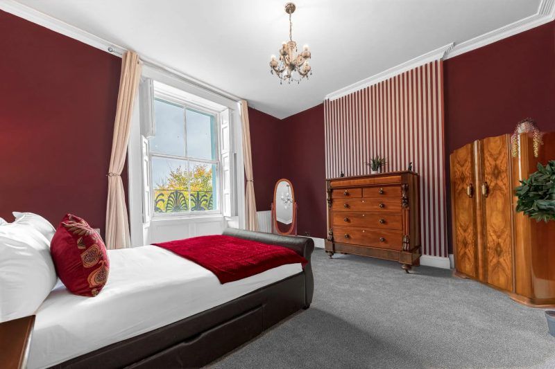 Bedroom with burgundy walls, gray carpet, and antique furniture; a large window with white trim.