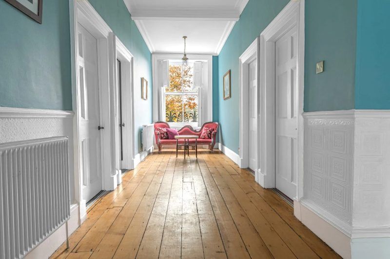 Long hallway with wooden floors, blue walls, white trim and doors, leading to a window with seating.