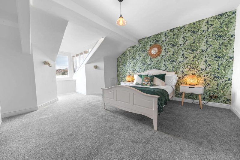 Bedroom with white bed, green patterned wallpaper, gray carpet, and a staircase.