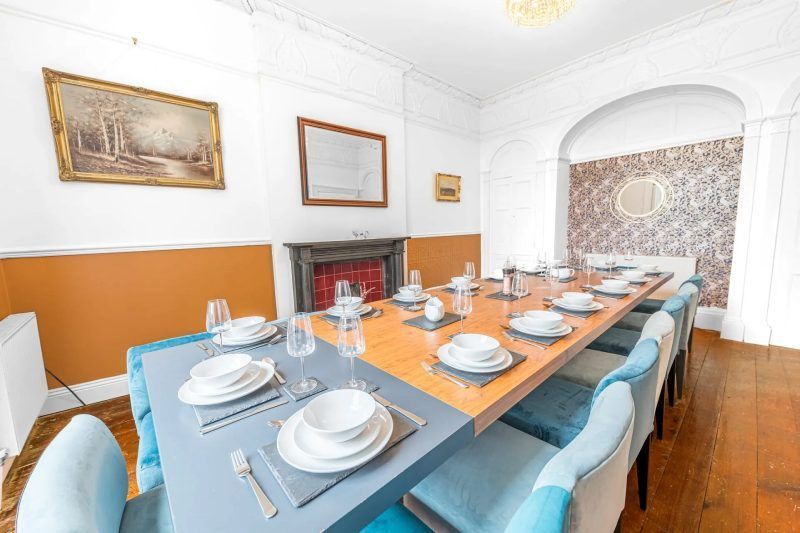 Dining room with long table set for many guests, blue chairs, wood floor, fireplace, and artwork.