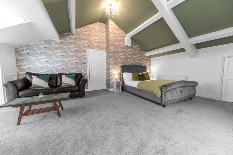 Bedroom with grey carpet, sofa, bed, wallpaper accent wall, and exposed beams.