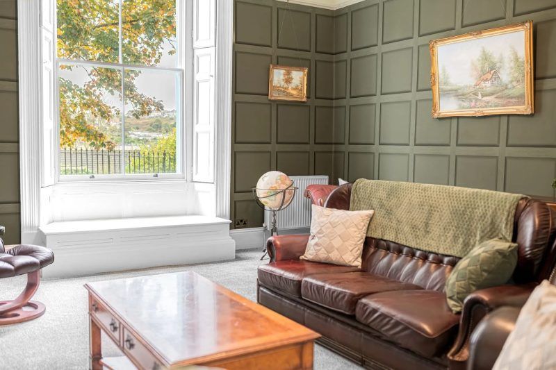 Living room with brown leather sofa, wood paneling, large window, artwork, and coffee table.