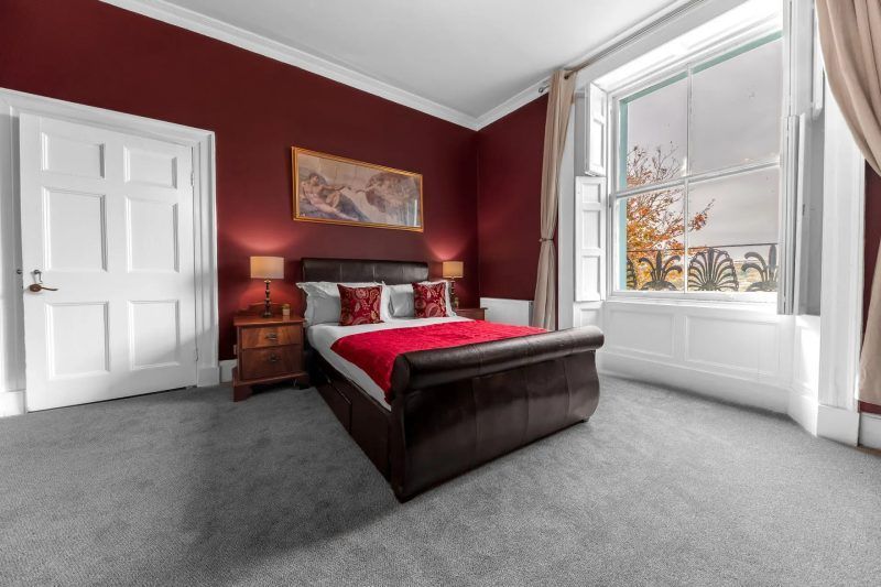 Burgundy bedroom with sleigh bed, framed art, nightstands, large window with foliage view, and white trim.
