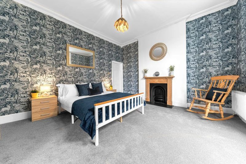 Bedroom with patterned wallpaper, white bed, fireplace, and rocking chair.