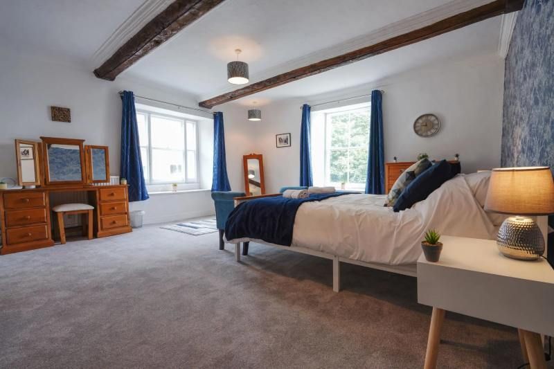Bedroom with a bed, vanity, and windows, featuring blue and white decor, dark wood beams, and grey carpet.