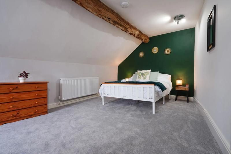 Bedroom with white bed, green accent wall, wooden dresser, grey carpet, and exposed beam.