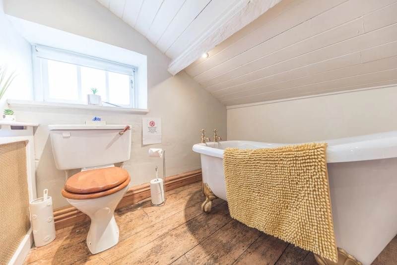 Bathroom with a white clawfoot bathtub, wooden floor, toilet, and small window. Yellow rug on the tub.