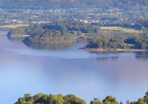 Calm lake reflects trees and sky, distant town visible.
