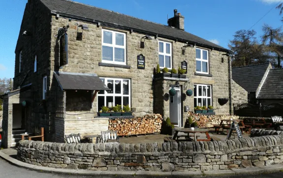 Stone pub with white-framed windows, wood stacks, and outdoor seating on a sunny day.