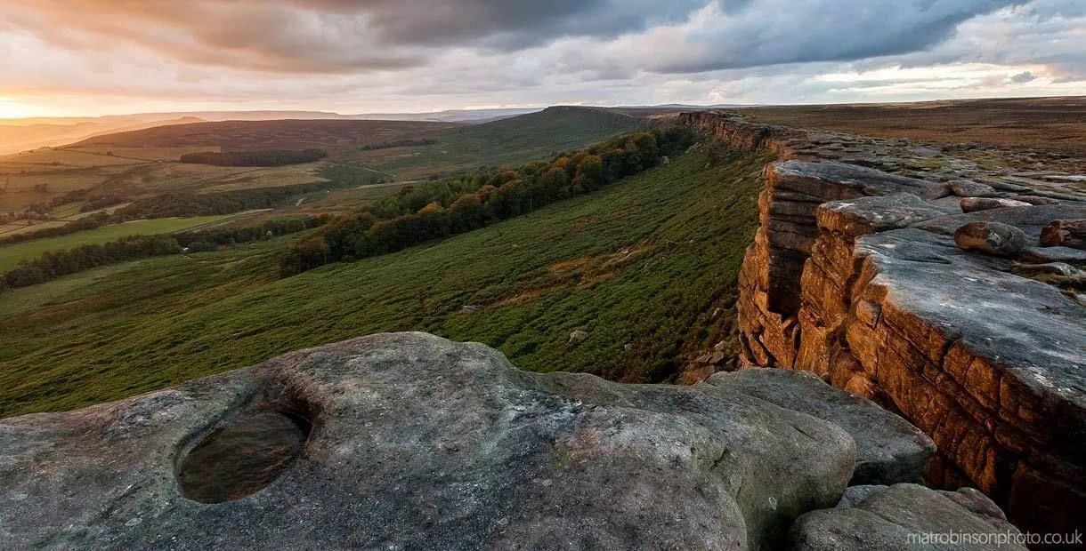 Dramatic cliff edge overlooking a valley with dark green trees. Cloudy sky with a sunrise glow.