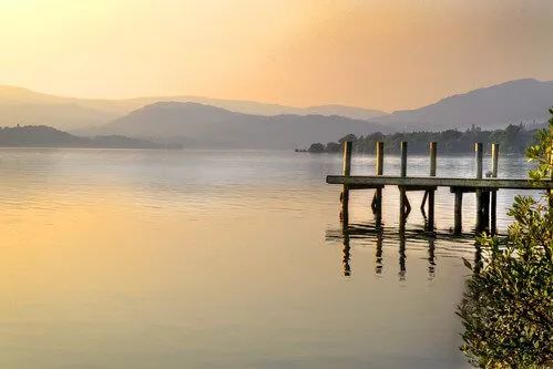 Calm lake scene at dusk with wooden pier and misty mountains in background.