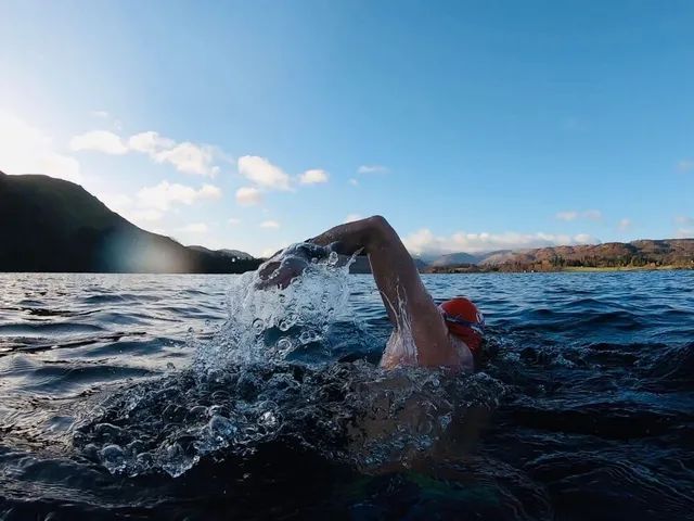 Person swimming freestyle stroke in a dark lake with mountains and blue sky in the background.