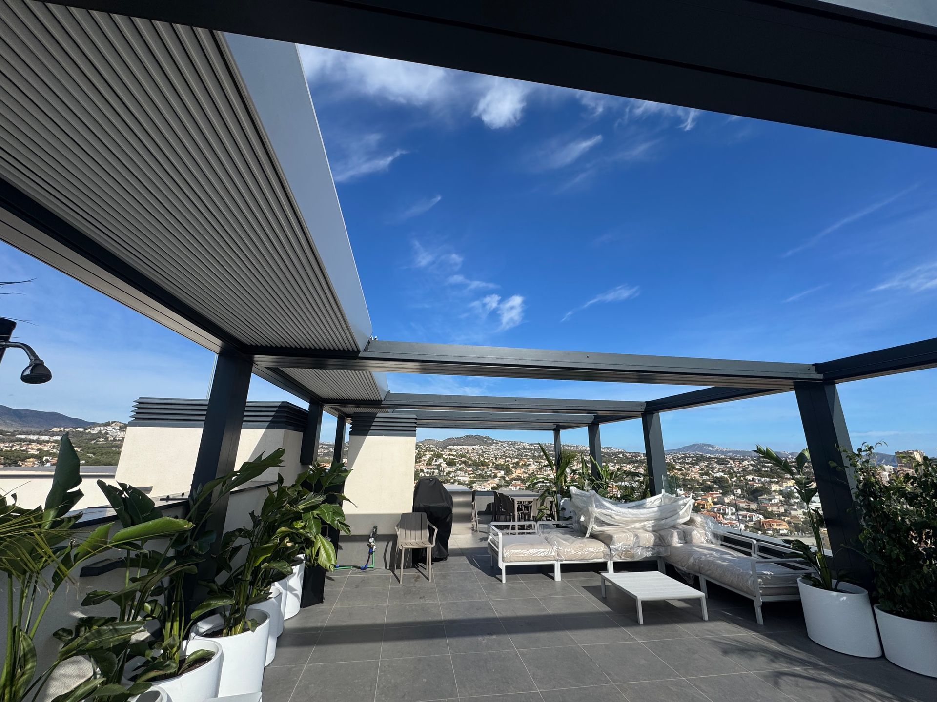 Patio en la azotea con techo retráctil, zona de estar y plantas en macetas, con vista a un paisaje urbano bajo un cielo azul.